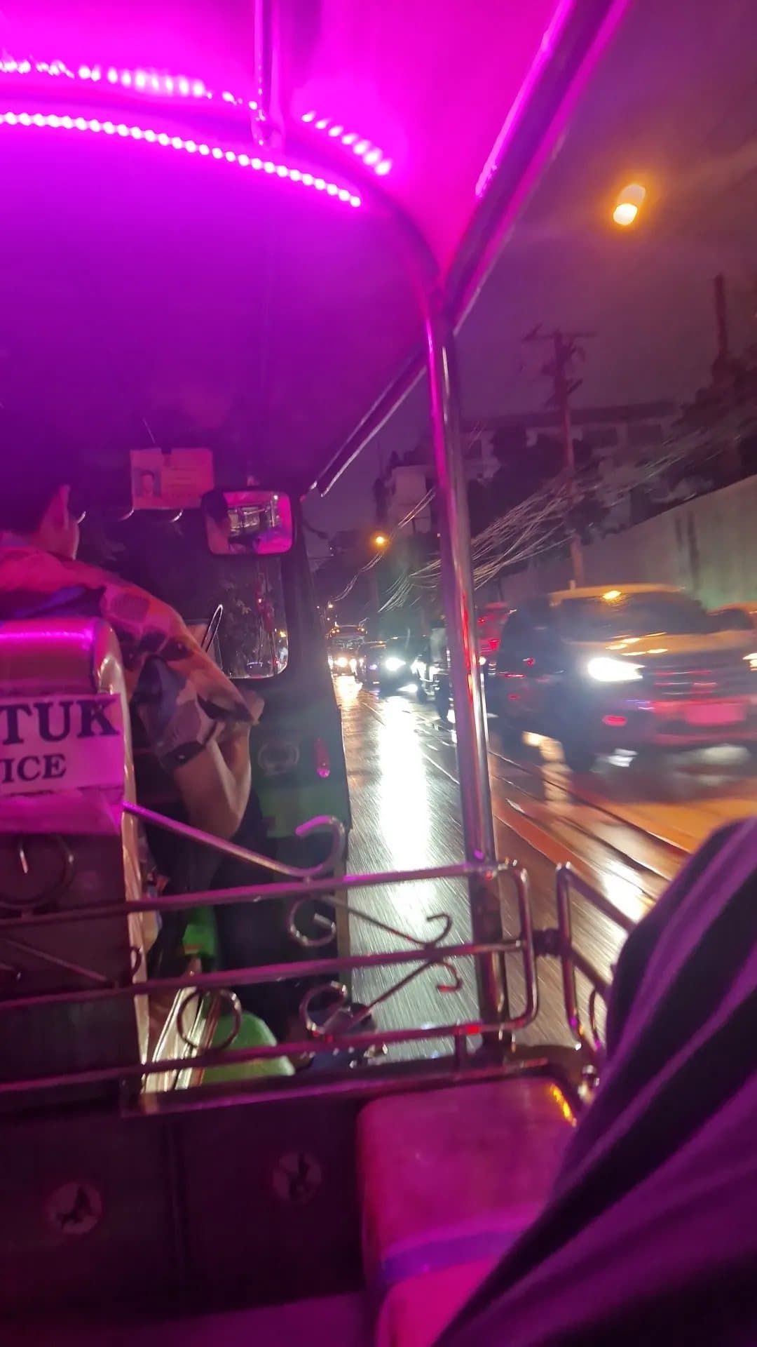 View from the back seat of a pink-lit tuk-tuk driving along a wet Bangkok road at night