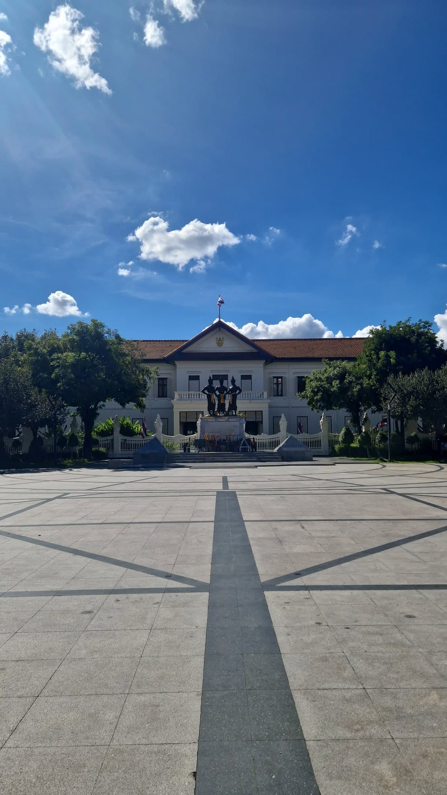 A wide paved plaza in front of a colonial-style building with a statue, under strong blue sky