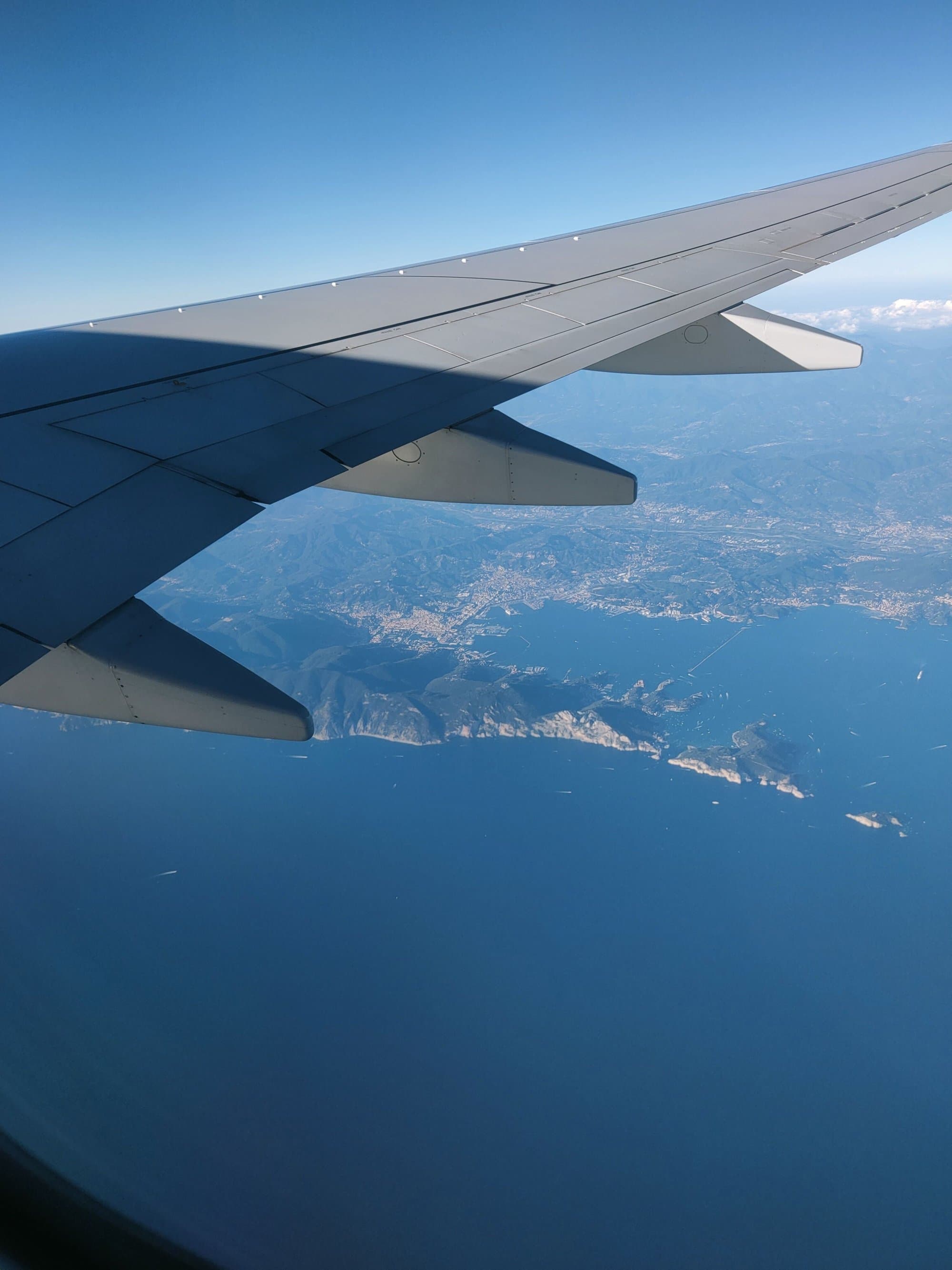 An aircraft wing over a blue sea and a rocky coastline, seen from a plane window